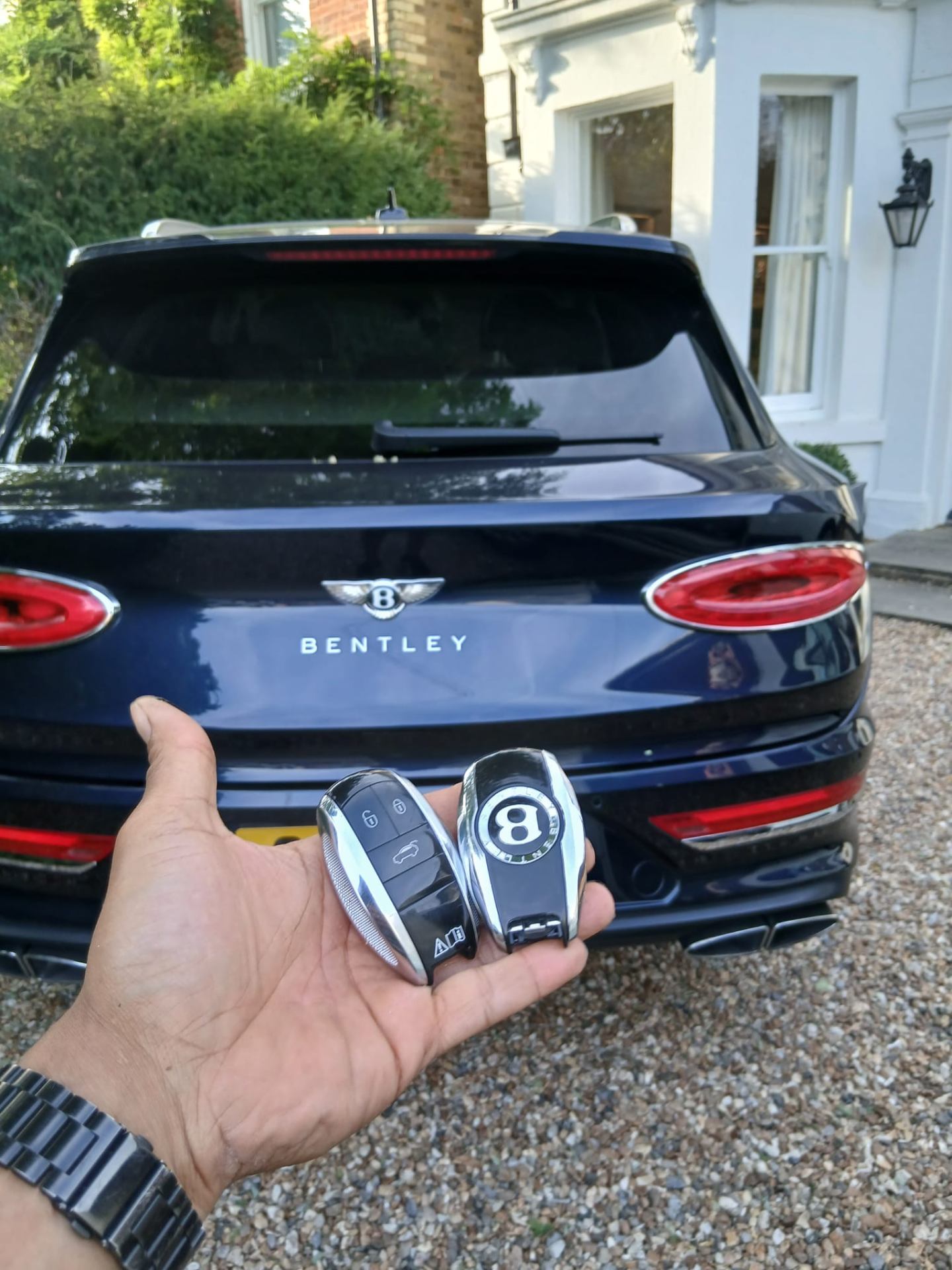 Two Bentley Bentayga smart keys held behind a dark blue Bentley Bentayga on a gravel driveway