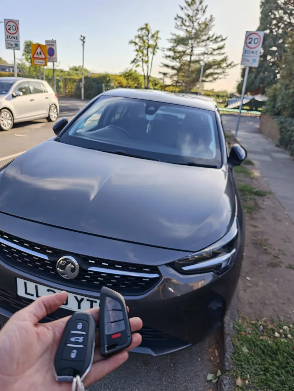 Two smart key fobs held in front of a grey Vauxhall Corsa parked at the kerb