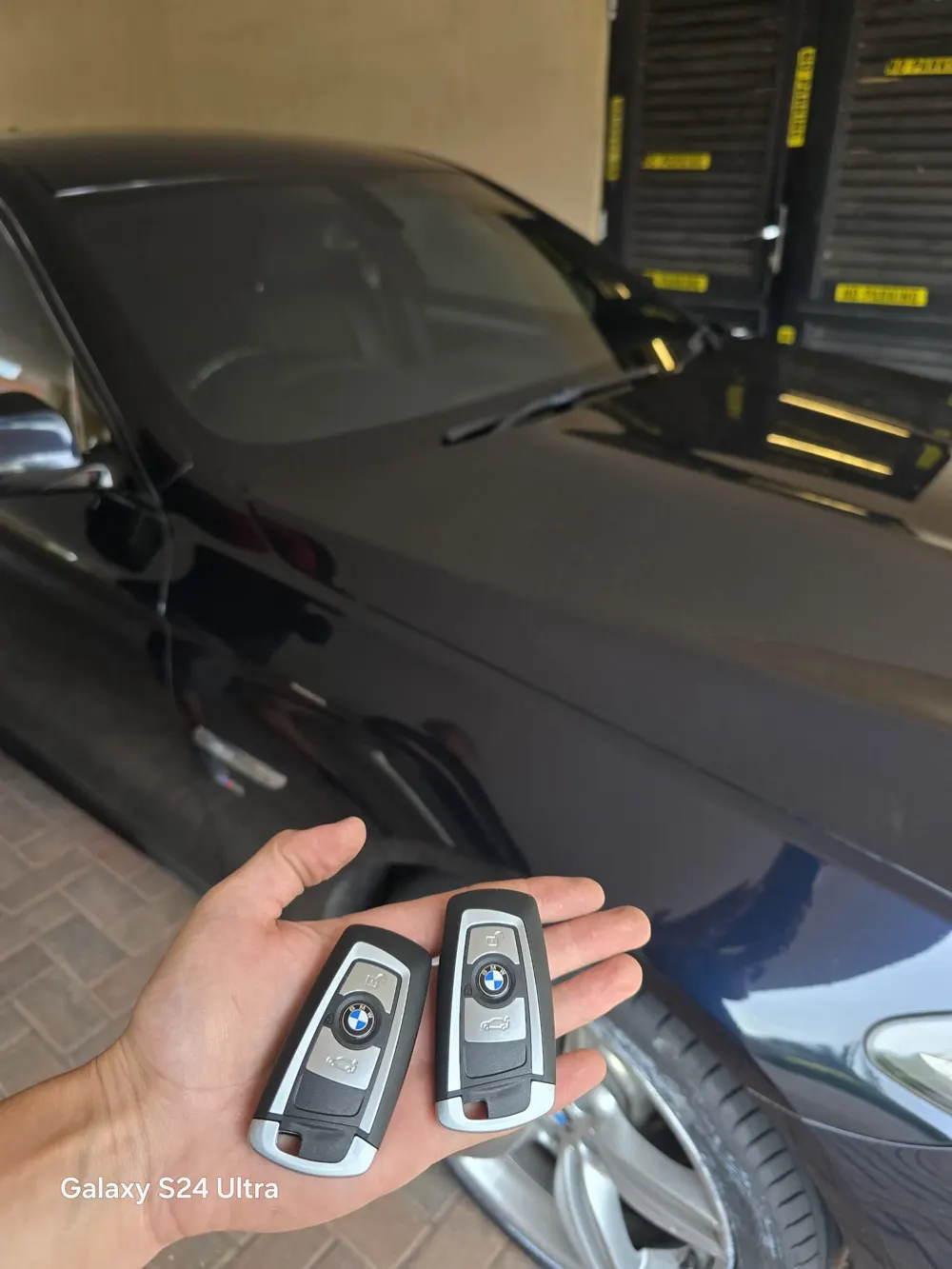 Two new BMW smart keys held beside a dark BMW parked on a driveway