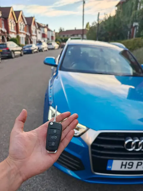 Replacement Audi flip key held up in front of a blue Audi Q3 on a residential street