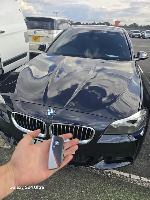 New BMW key fob held in front of a dark blue BMW 5 Series in a car park